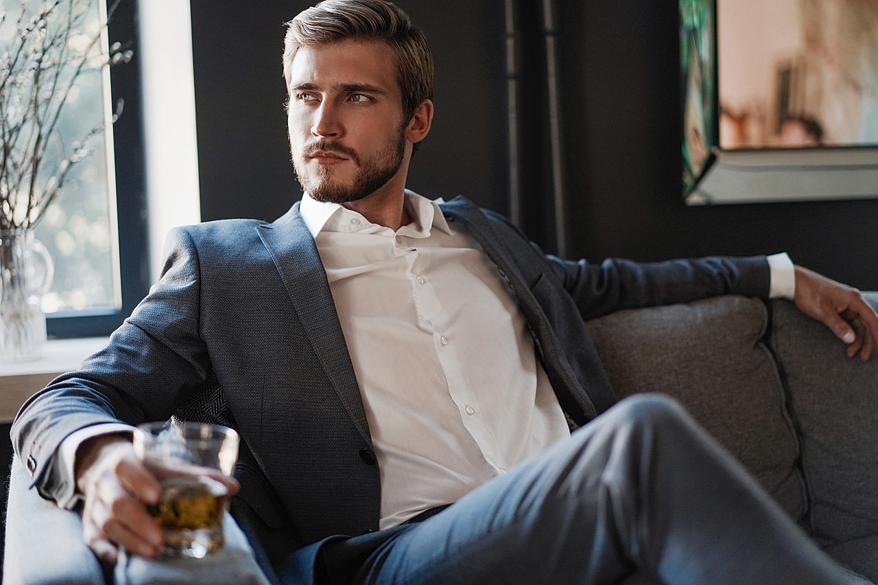 Man in suit sipping whiskey while sitting.